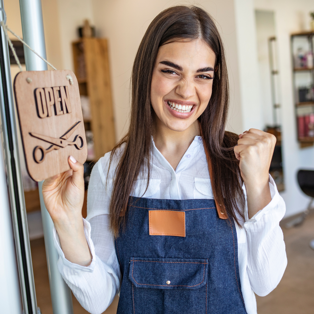 Brunette woman wearing an apron and clasping her fist in excitement while flipping her business door sign to "open". Gives the appearance she is celebrating the first day of commerce as a business owner.