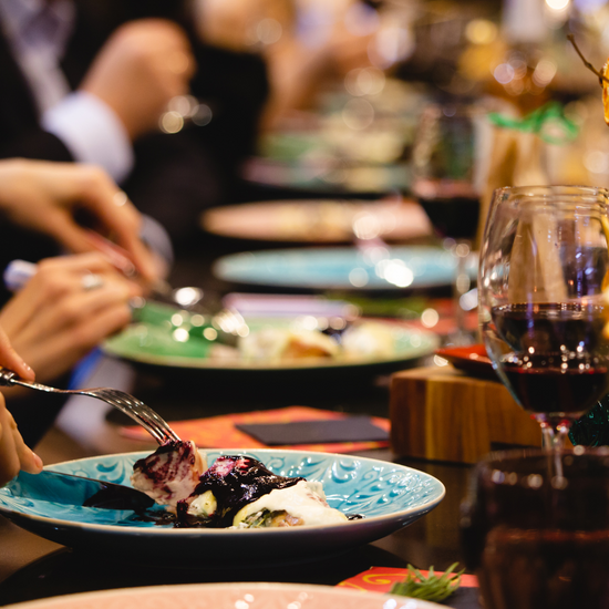 Long, wooden, fine-dining dinner table, decorated with wooden accents and colorful plates, topped with gourmet food, and accompanied by wine glasses topped with dark, red wine. The photo focuses on the hands of the diners as they enjoy their meal.