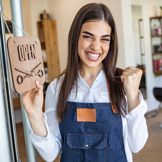 Brunette woman wearing an apron and clasping her fist in excitement while flipping her business door sign to 